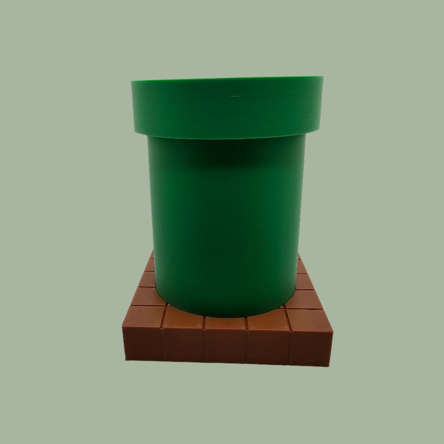 A 3D-printed pen holder designed to resemble a green pipe from a classic video game, sitting on a brown, block-patterned base. The cylindrical holder is vibrant green with a slightly wider rim at the top. The brown base mimics the look of pixelated bricks.