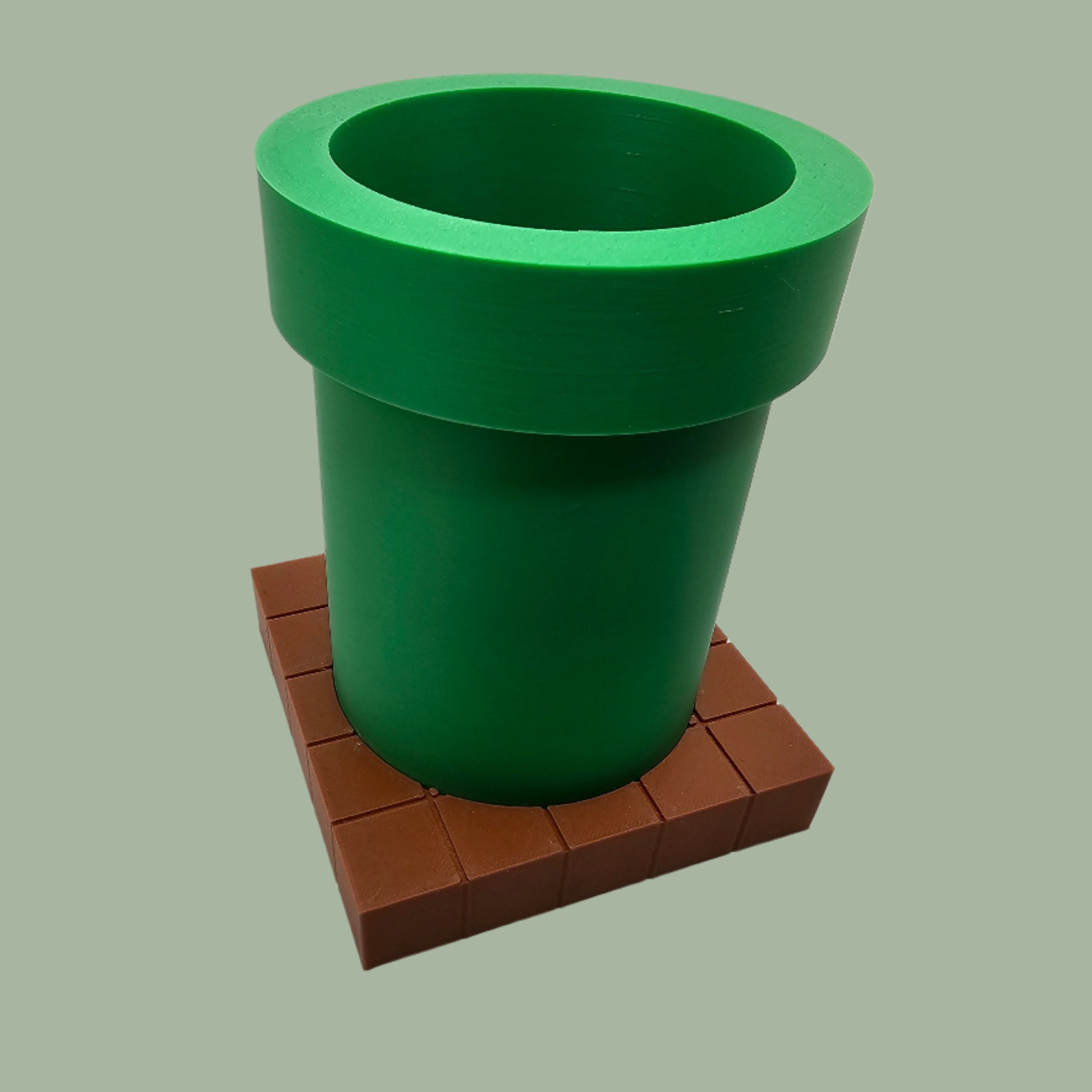 A 3D-printed pen holder designed to resemble a green pipe from a classic video game, sitting on a brown, block-patterned base. The cylindrical holder is vibrant green with a slightly wider rim at the top. The brown base mimics the look of pixelated bricks.