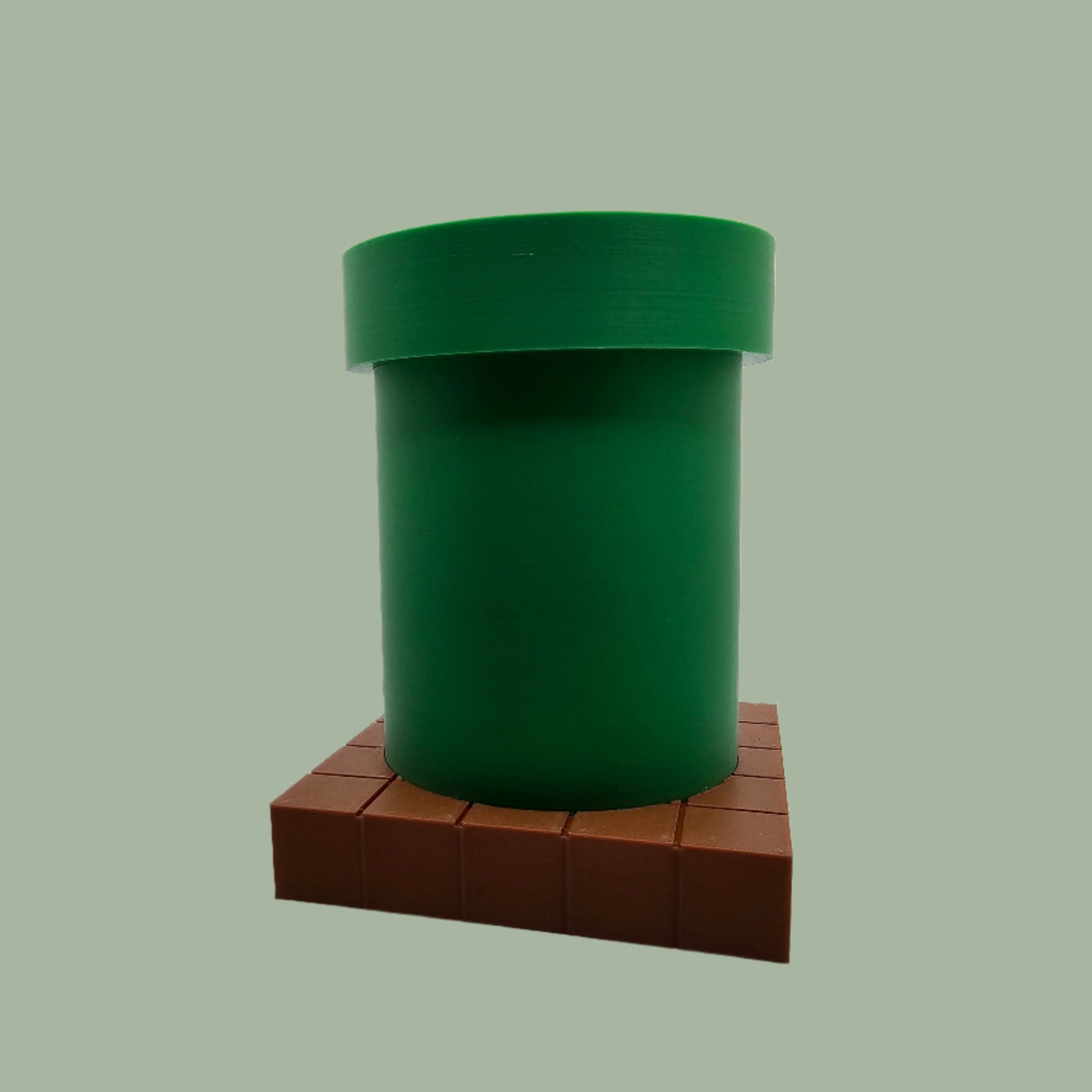 A 3D-printed pen holder designed to resemble a green pipe from a classic video game, sitting on a brown, block-patterned base. The cylindrical holder is vibrant green with a slightly wider rim at the top. The brown base mimics the look of pixelated bricks.