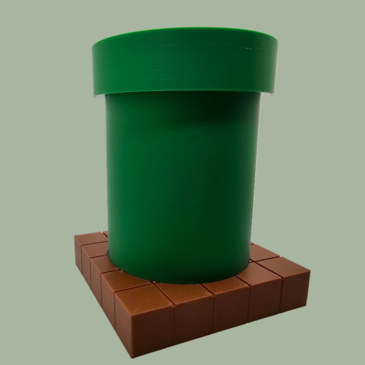 A 3D-printed pen holder designed to resemble a green pipe from a classic video game, sitting on a brown, block-patterned base. The cylindrical holder is vibrant green with a slightly wider rim at the top. The brown base mimics the look of pixelated bricks.