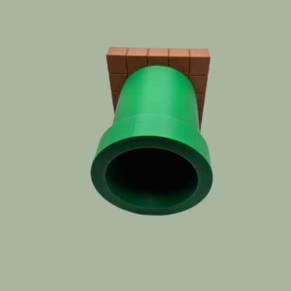 A 3D-printed pen holder designed to resemble a green pipe from a classic video game, sitting on a brown, block-patterned base. The cylindrical holder is vibrant green with a slightly wider rim at the top. The brown base mimics the look of pixelated bricks.