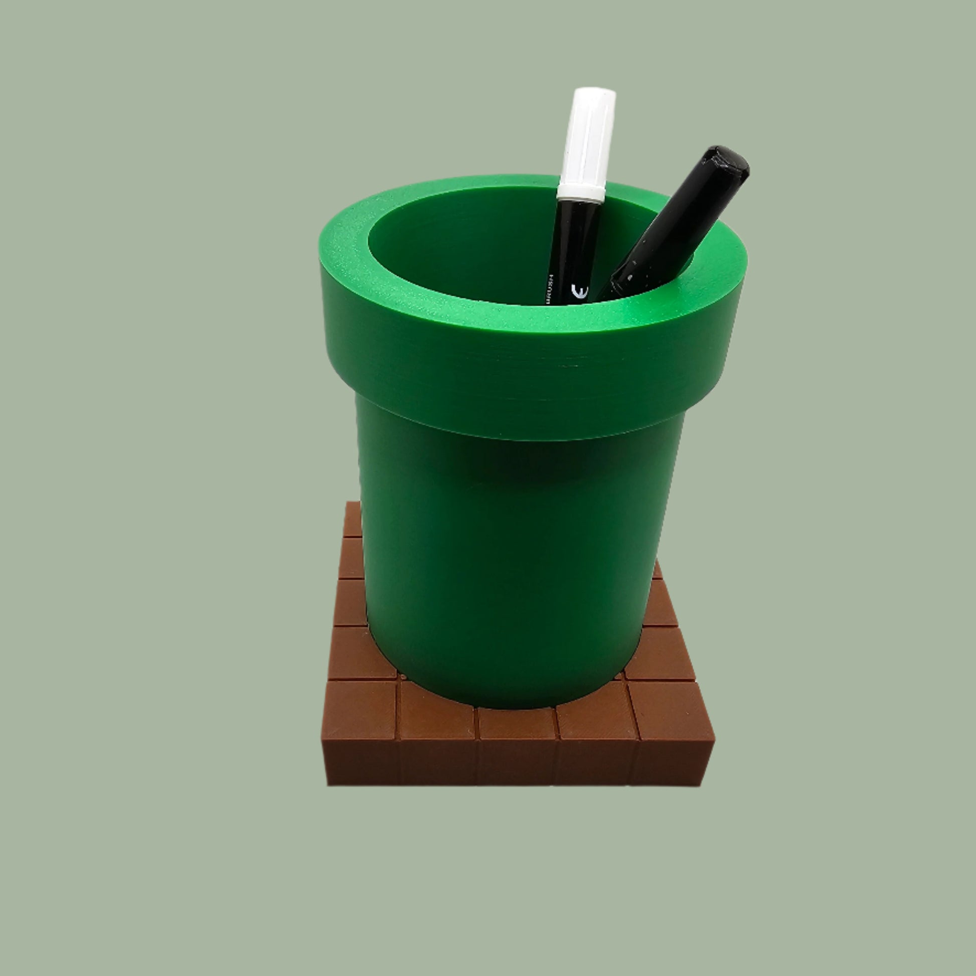 A 3D-printed pen holder designed to resemble a green pipe from a classic video game, sitting on a brown, block-patterned base. The cylindrical holder is vibrant green with a slightly wider rim at the top. The brown base mimics the look of pixelated bricks.