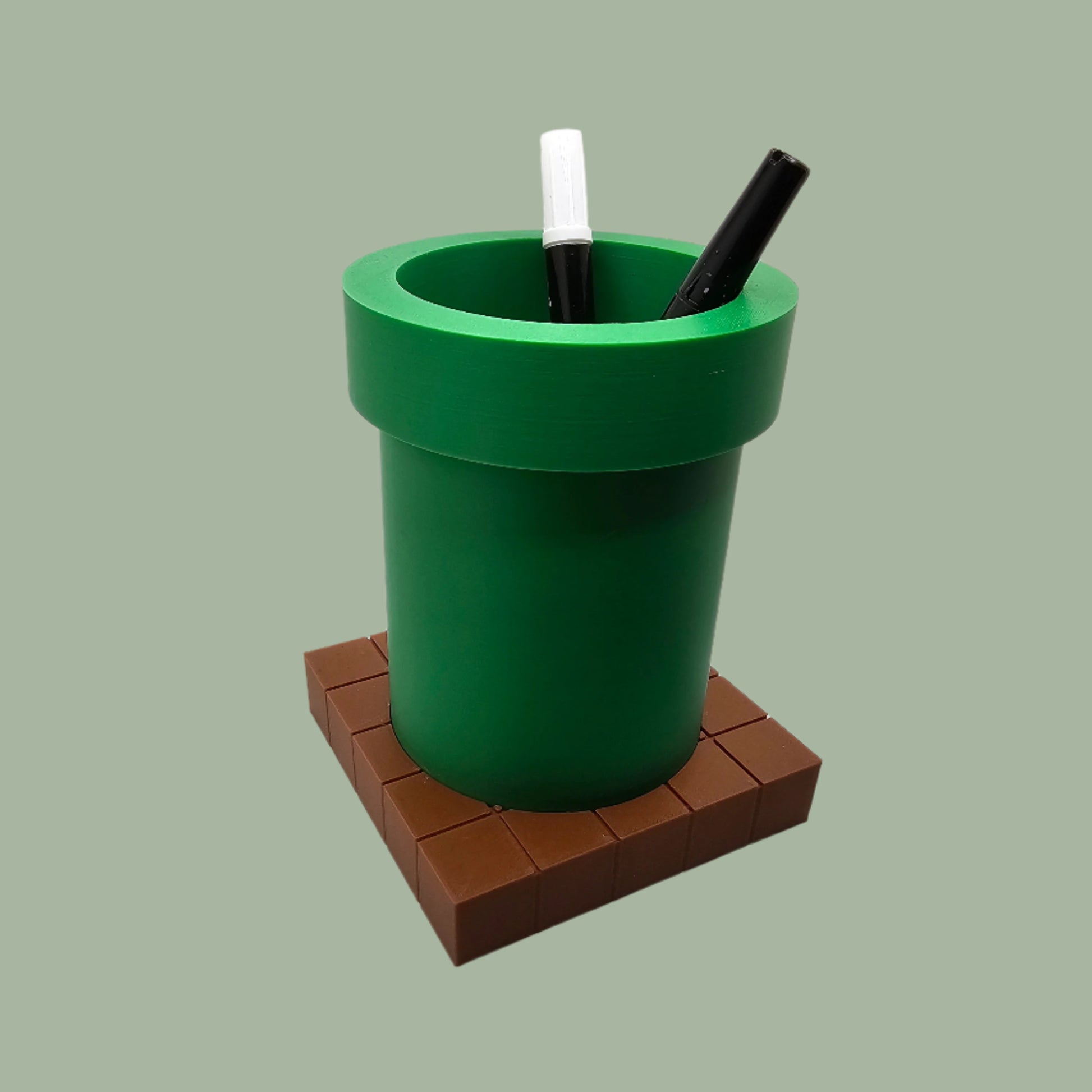 A 3D-printed pen holder designed to resemble a green pipe from a classic video game, sitting on a brown, block-patterned base. The cylindrical holder is vibrant green with a slightly wider rim at the top. The brown base mimics the look of pixelated bricks.